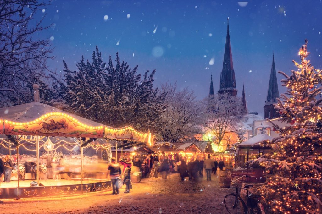 A traditional German Christmas market scene under a starry sky. There is a Christmas tree in the foreground and a church behind the market.